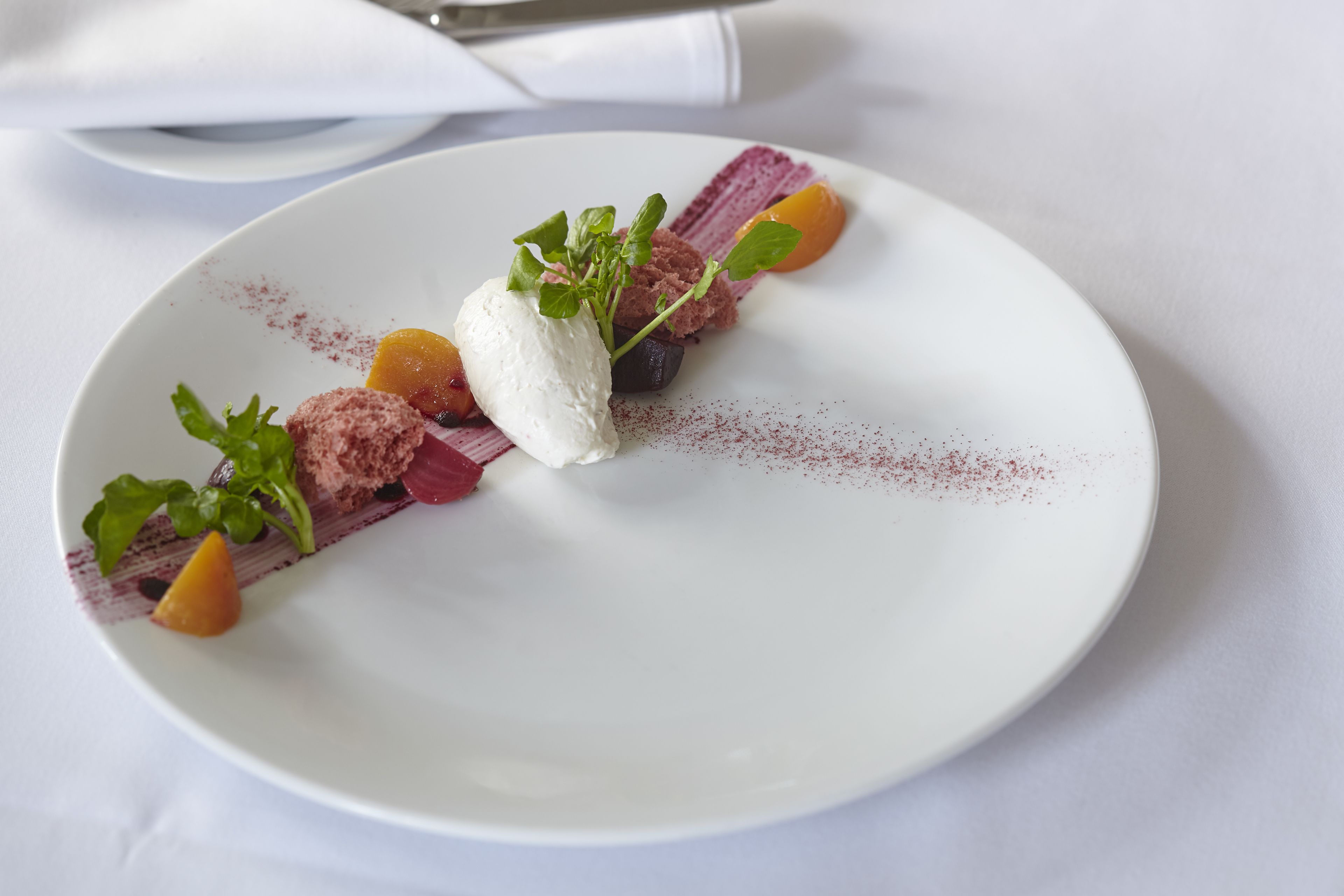 A white plate with a dessert consisting of a scoop of ice cream, a piece of fruit, and some greens.