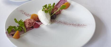 A white plate with a dessert consisting of a scoop of ice cream, a piece of fruit, and some greens.