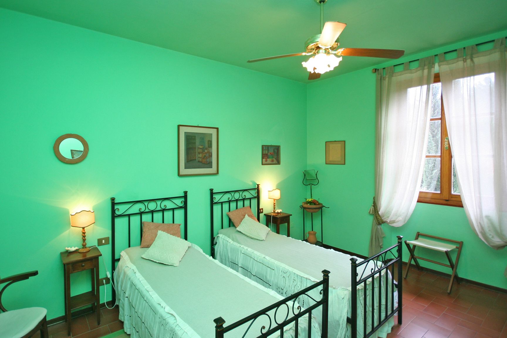 A room with two beds, a ceiling fan, a window with curtains, and a small table with a lamp.