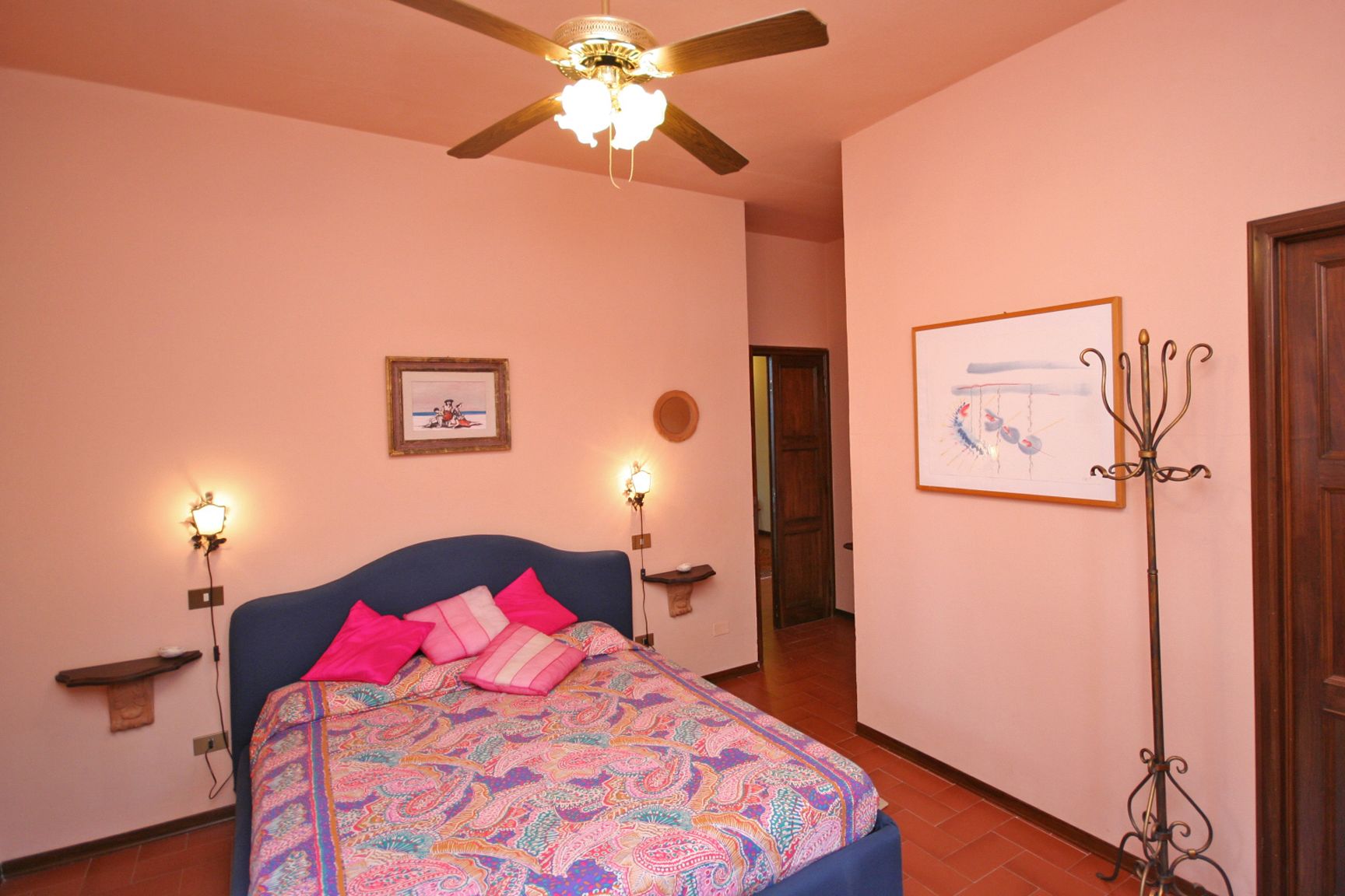 A bedroom with a bed, a ceiling fan, wall-mounted lights, a framed picture, and a floor lamp.