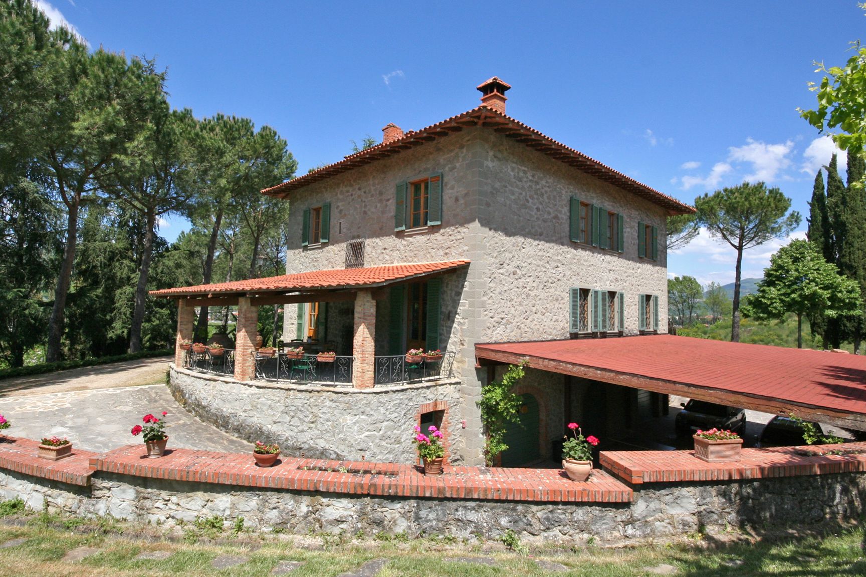 Una casa de piedra con techo de tejas rojas, un balcón con mobiliario exterior y un muro de piedra que rodea la propiedad.