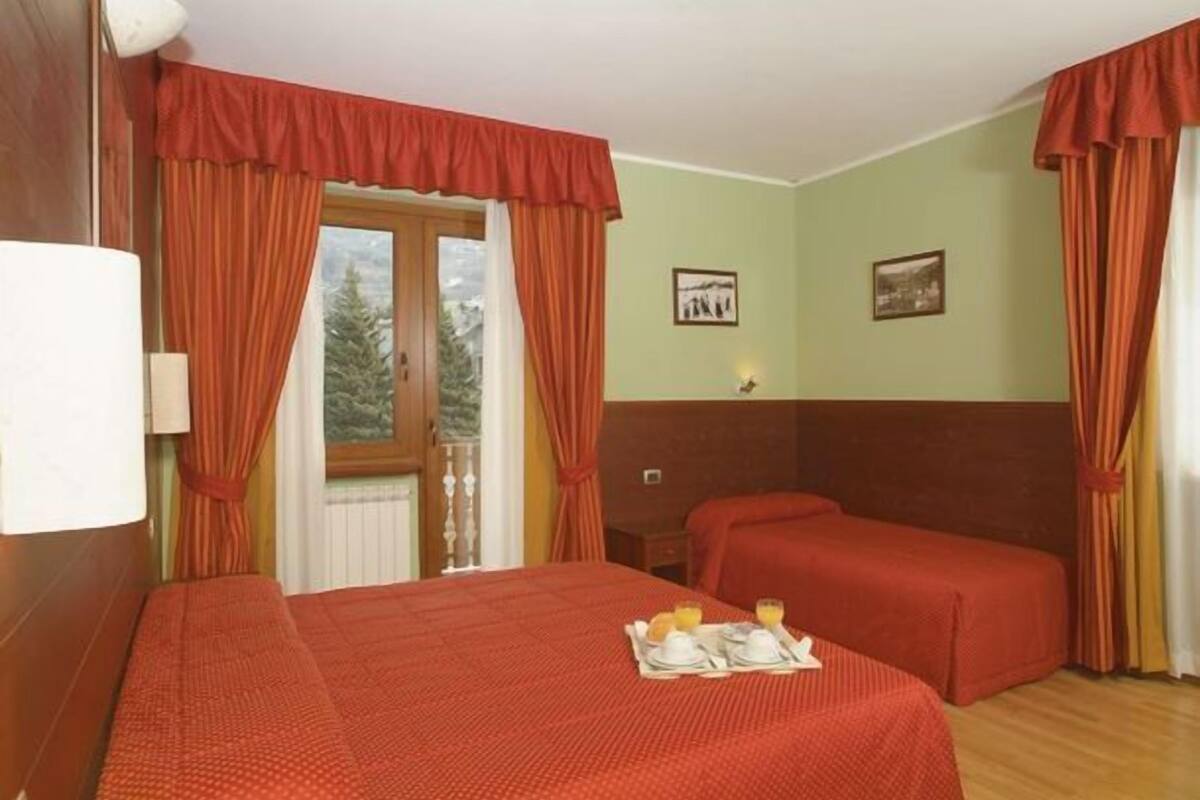 Ein Hotelzimmer mit zwei Einzelbetten, einem Fenster mit Vorhängen, einem kleinen Tisch mit Frühstücksgebäck und zwei gerahmten Bildern an der Wand.