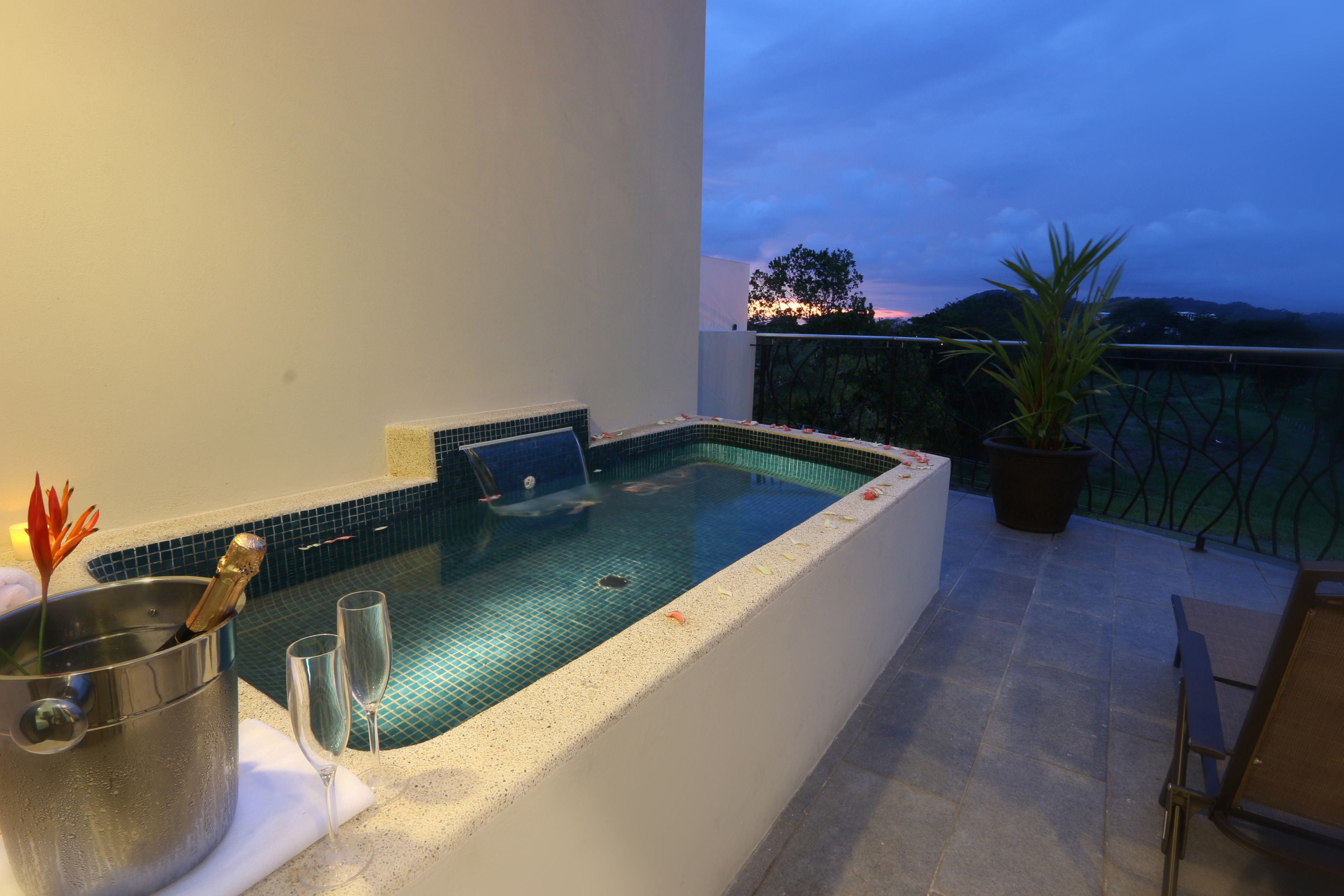 A hot tub with a tiled interior, a potted plant, and a glass of champagne on a balcony with a view of the sunset.