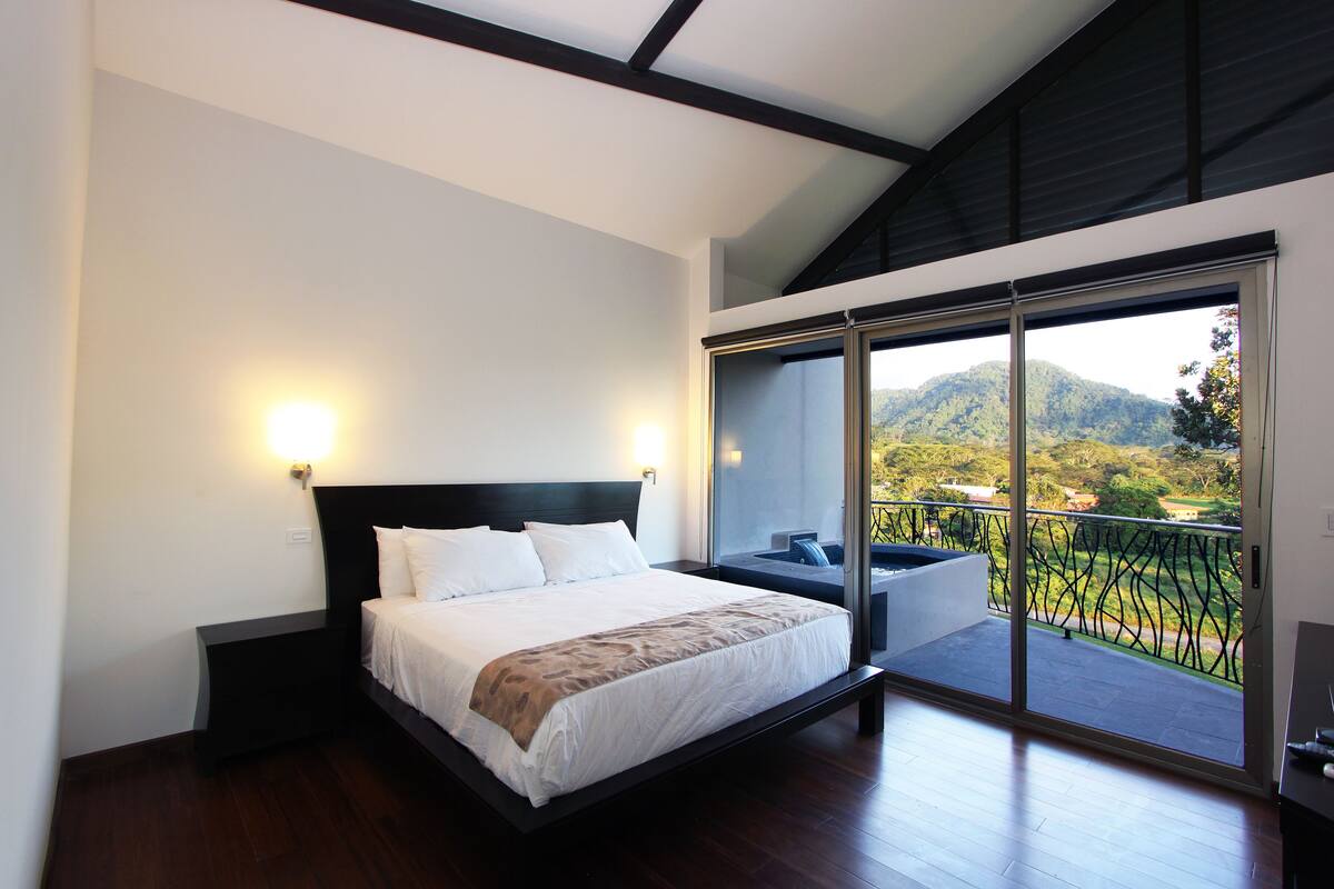 A modern bedroom with a large bed, a balcony with a view of the mountains, and a sliding glass door.