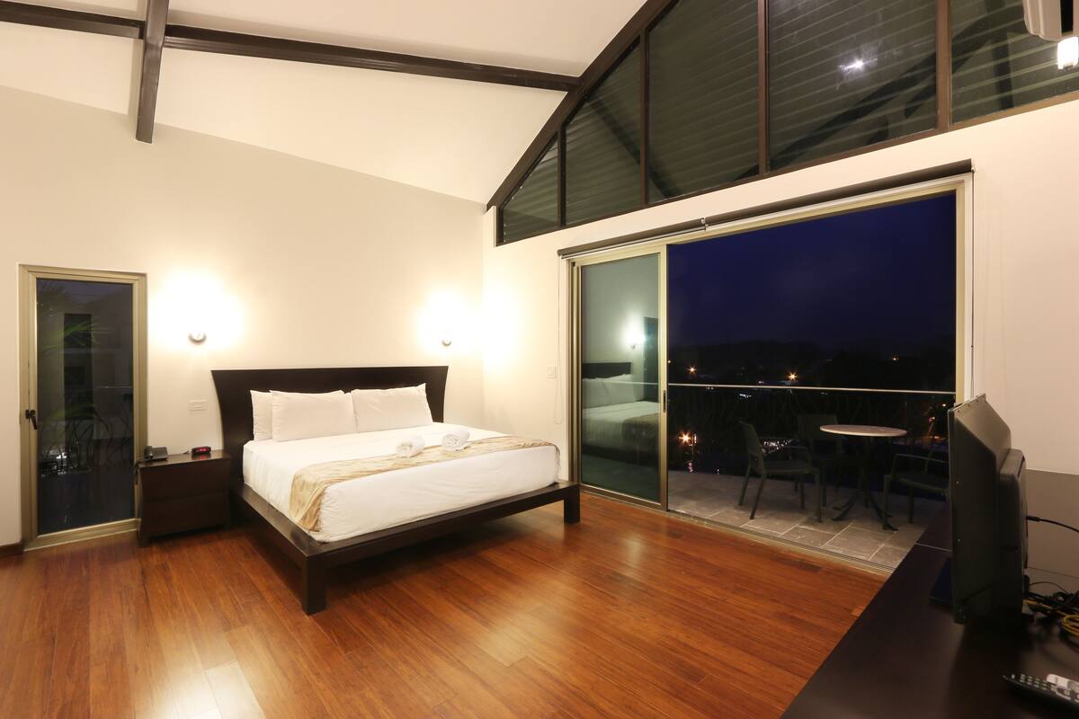 A bedroom with a bed, bedside tables, a TV, and a balcony with a table and chairs.