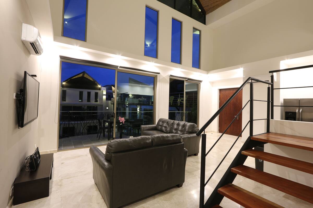 A spacious living area with a modern staircase, a large window overlooking a cityscape, and a comfortable seating arrangement.