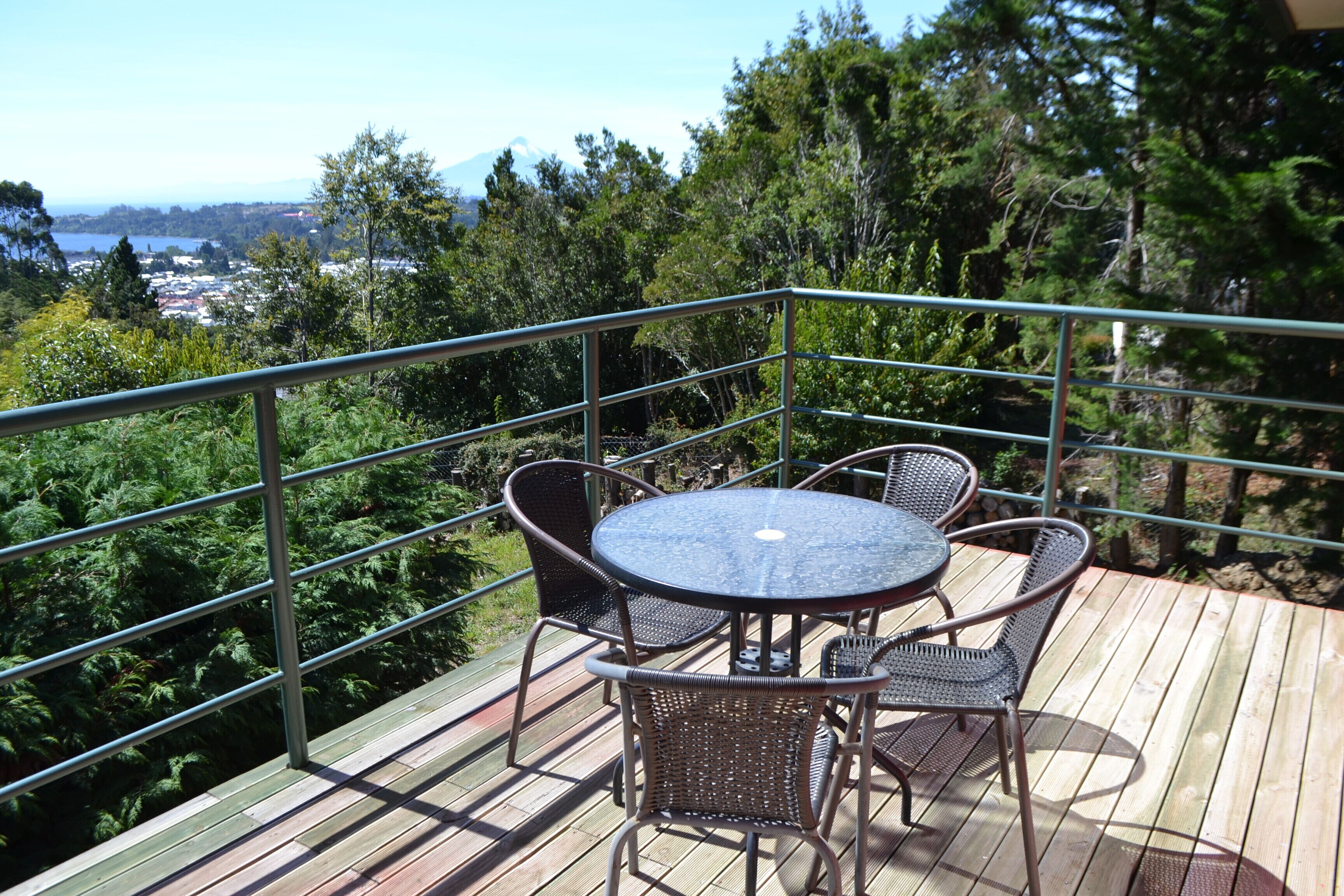 A wooden deck with a round table and four chairs, surrounded by a metal railing, overlooking a forested area with a distant view of a town and a mountain.