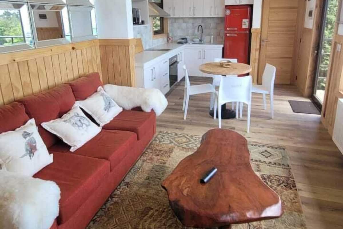 Una sala de estar moderna con un sofá rojo, una mesa de centro de madera y una alfombra estampada. Hay una cocina con armarios blancos y un refrigerador rojo.