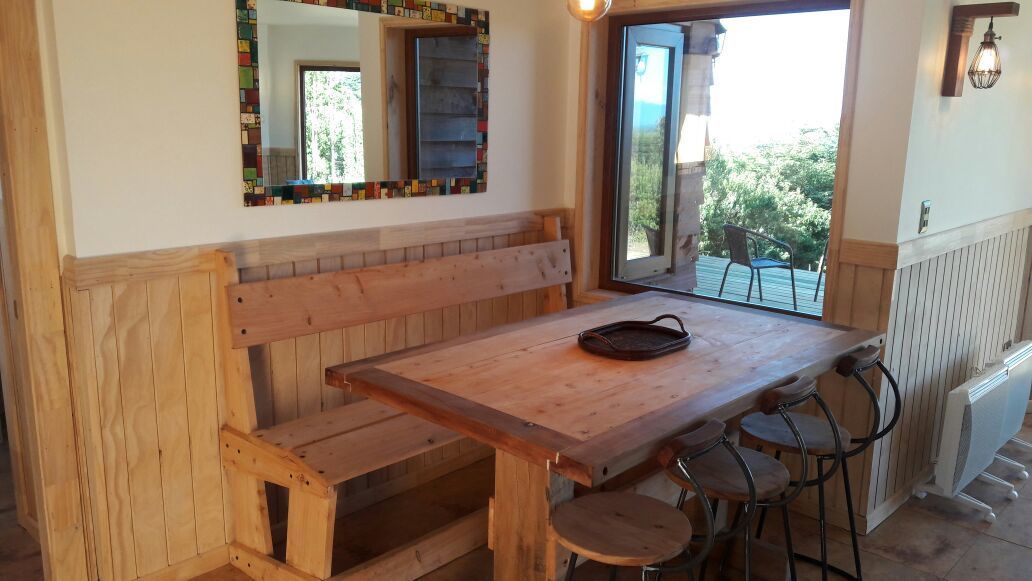 Un banco y una mesa de madera con un bolso negro encima, un espejo con un marco colorido, y vistas al exterior con vegetación.