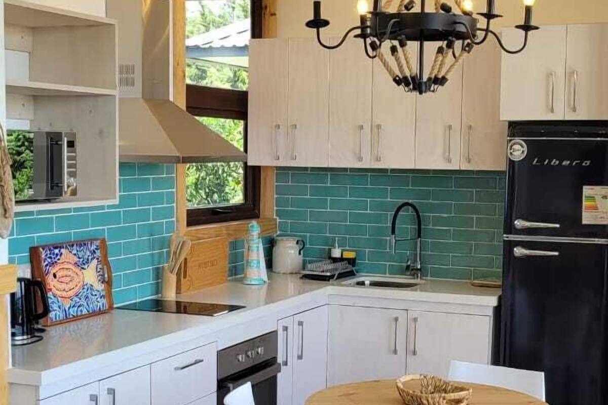 Una cocina moderna con armarios blancos, un refrigerador negro, una mesa de comedor de madera con sillas blancas y una araña.
