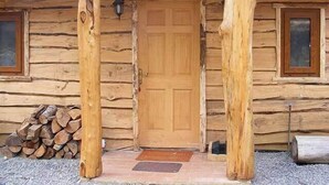 Una cabaña de troncos con puerta de madera, dos ventanas y leña apilada afuera.
