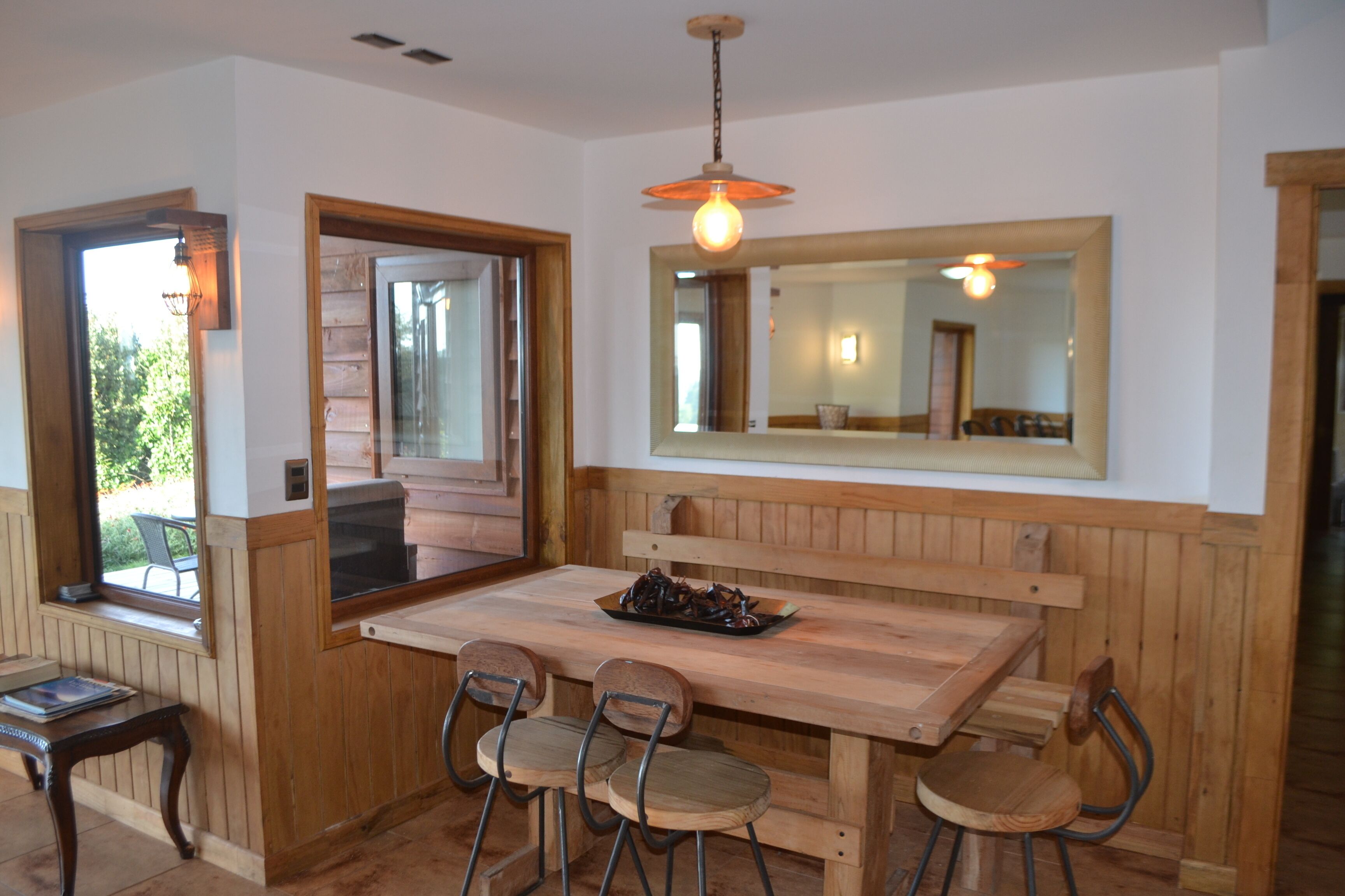 Une table à manger en bois, accompagnée de quatre chaises, un luminaire suspendu et un grand miroir placé au-dessus de la table.