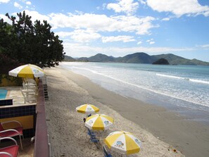 A beach with umbrellas and chairs, a pool, and mountains in the background. - Pousada Casa na Praia (Ubatuba)