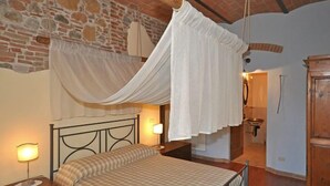 A bedroom with a bed, a nightstand, a lamp, a wooden wardrobe, and a stone wall. - Villa Cassia (Pienza)