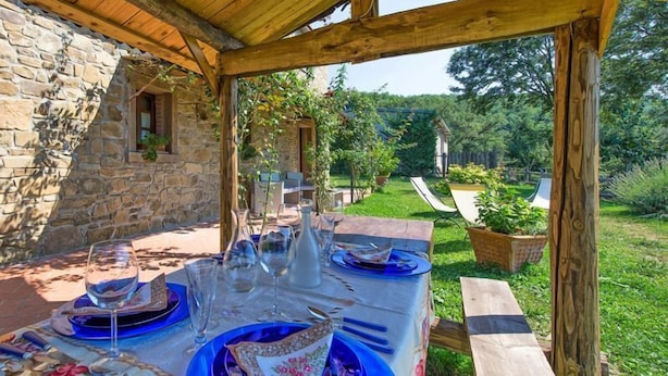An outdoor dining area with a stone wall, wooden pergola, and a view of a garden and house in the background. - Villa Margherita (Cortona)
