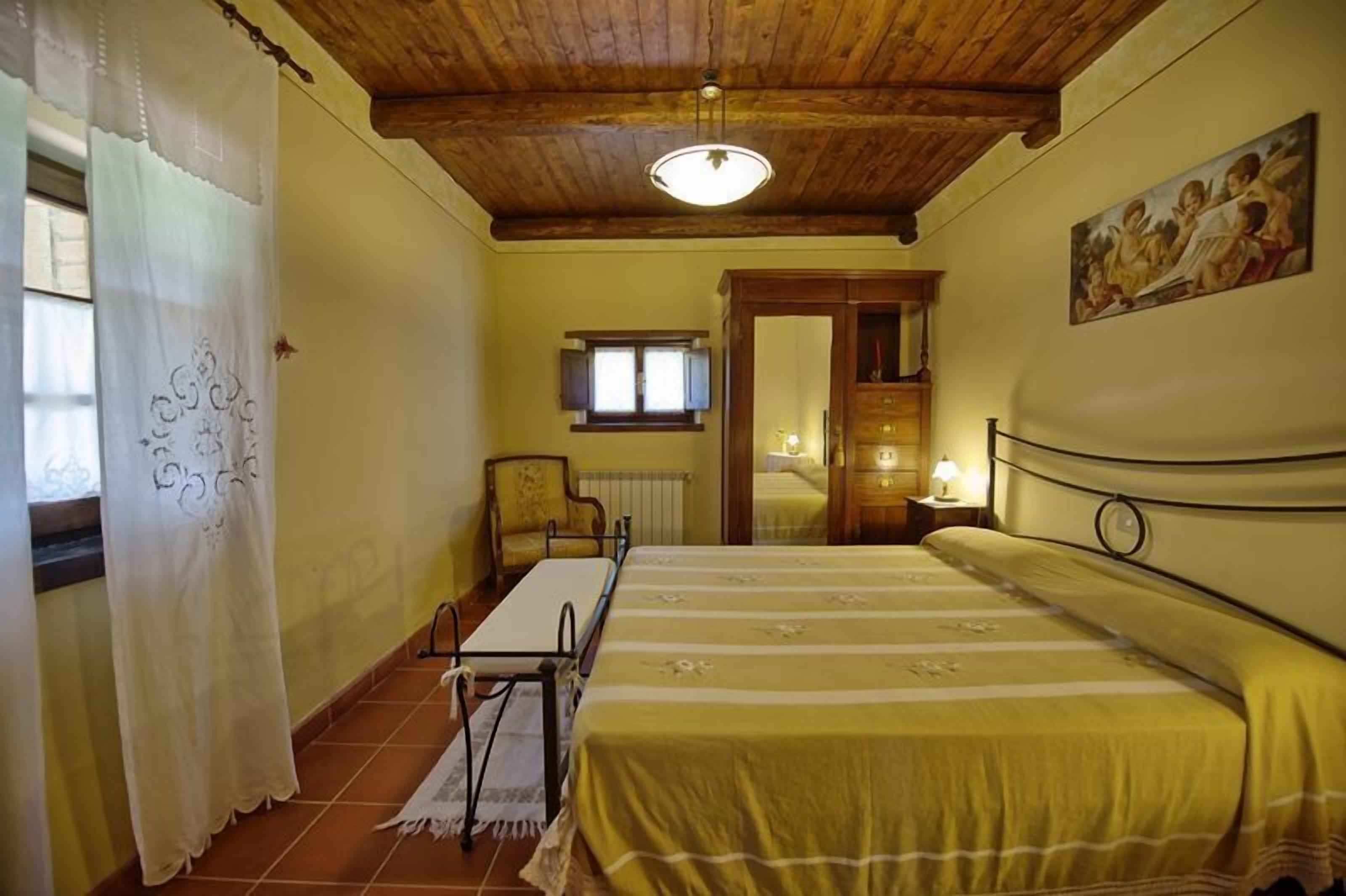 A room with a wooden ceiling, a bed with a yellow bedspread, a table with a chair, a window with a curtain, and a painting on the wall.