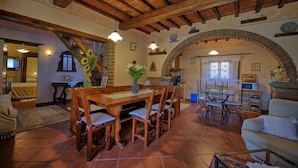 A rustic dining area with a wooden table and chairs, a stone archway, and a cozy living space with a sofa and a television. - Villa Margherita (Cortona)