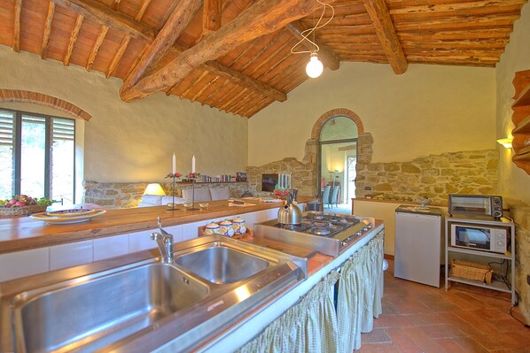 A rustic kitchen with a stone wall, wooden beams, a large sink, a stove, and a microwave. - Casa Uscioli (Pontassieve)