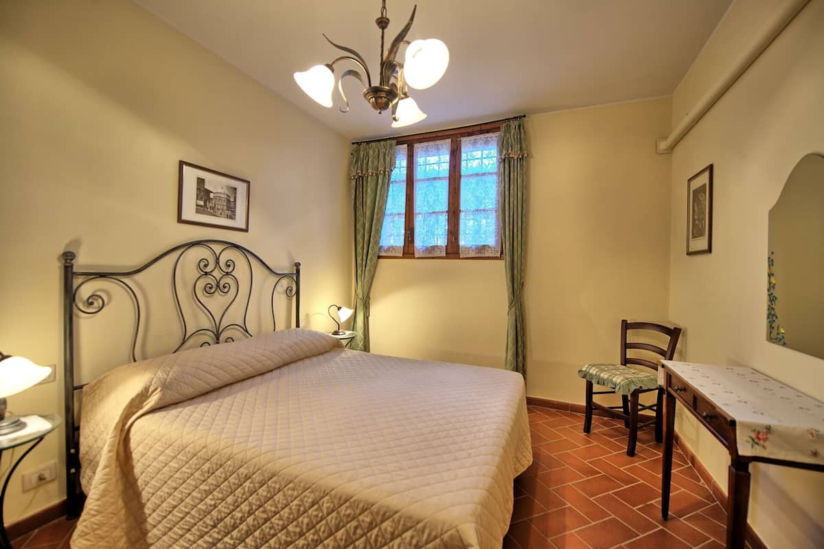 A bedroom with a bed, a chair, a desk, a window with curtains, and a chandelier.