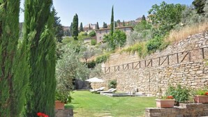A stone wall with a garden, a stone pathway, and a view of buildings in the background. - Villa Calcinaio (Cortona)