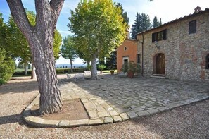 A stone building with a rustic exterior, a paved courtyard, and a large tree in the foreground. - Villa Guia (Bucine)