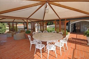 A covered outdoor patio with a round table and white chairs, surrounded by a wooden pergola structure. - Villa Guia (Bucine)