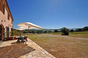 A patio with stone flooring, outdoor furniture, and umbrellas, overlooking a scenic landscape with hills and a body of water. - Talamone (Orbetello)