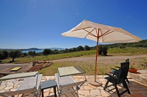 An outdoor seating area with tables and chairs, a large umbrella, and a scenic view of the countryside and a lake. - Talamone (Orbetello)