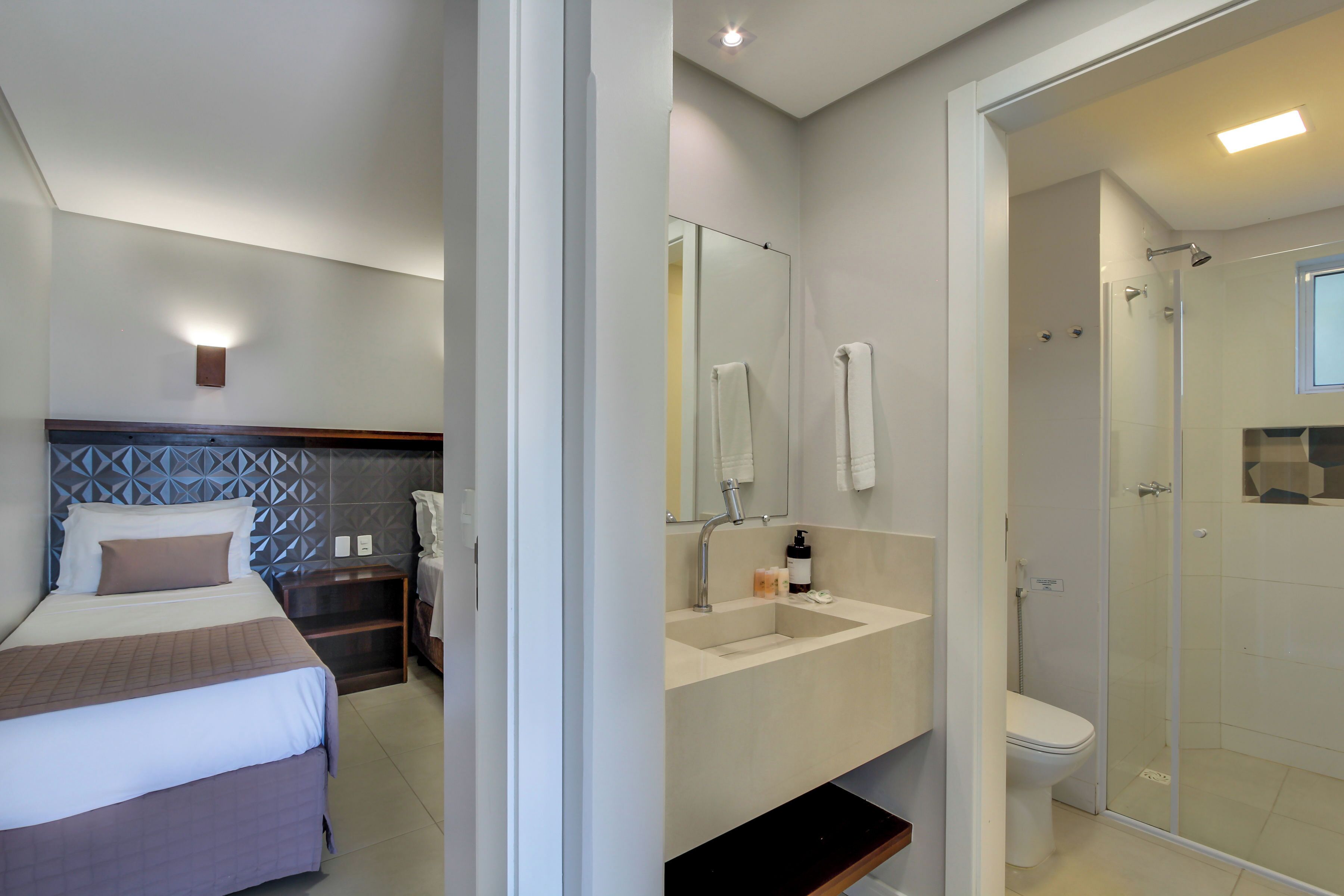 Quarto de hotel com cama, banheiro com pia, espelho e chuveiro.