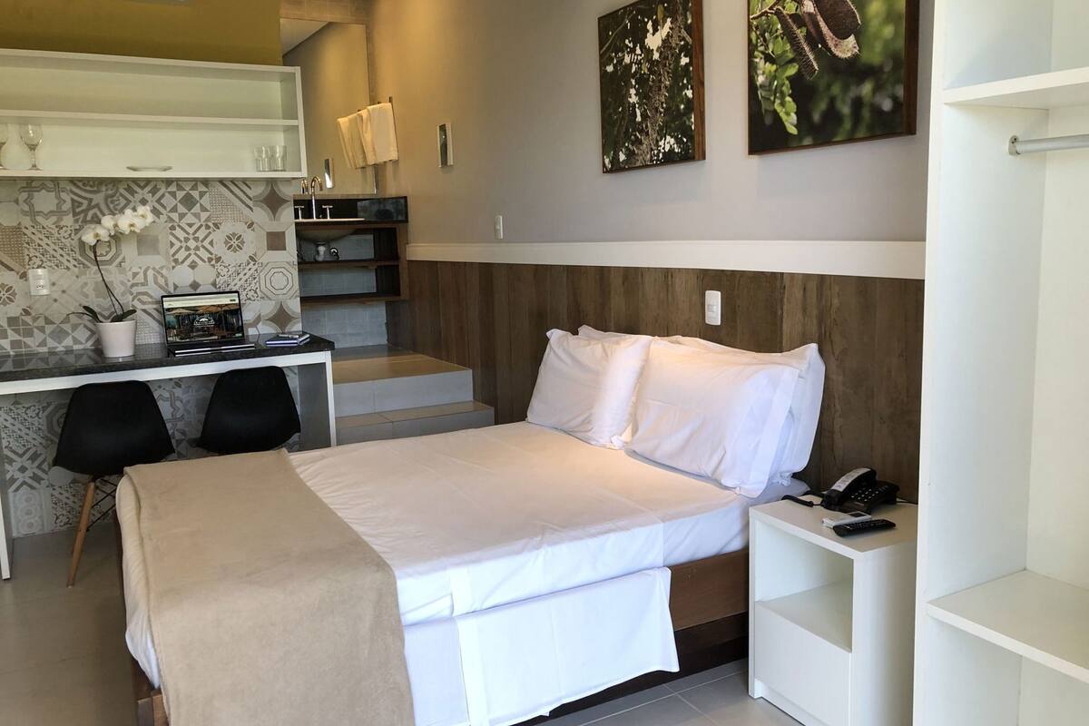 A modern hotel room with a bed, desk, and chair, a small kitchenette, and artwork on the walls.