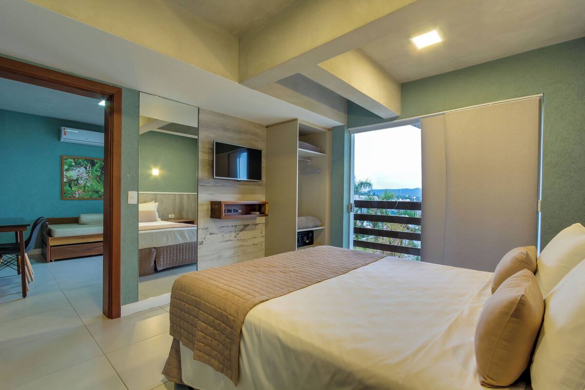 A bedroom with a bed, a TV, a balcony with a view, and a desk with a chair.
