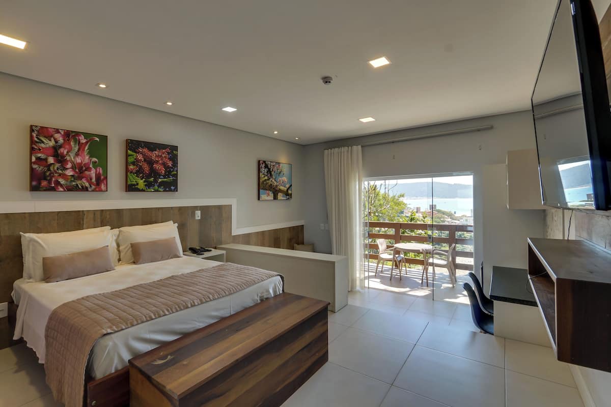 A modern hotel room with a large bed, a wooden bench, a desk, and a balcony with a view.