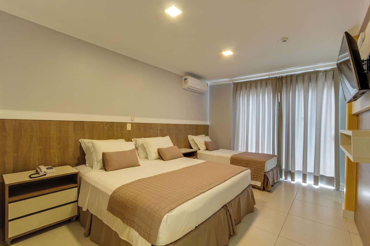 A hotel room with two beds, a TV, an air conditioning unit, a bedside table with a phone, and a window with curtains.