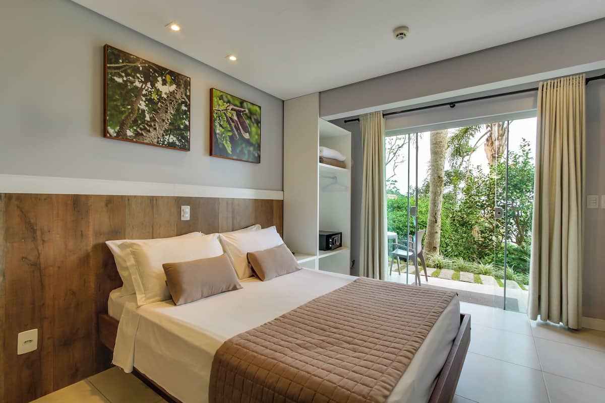 A modern bedroom with a large bed, wooden headboard, and a view of greenery through a sliding glass door.