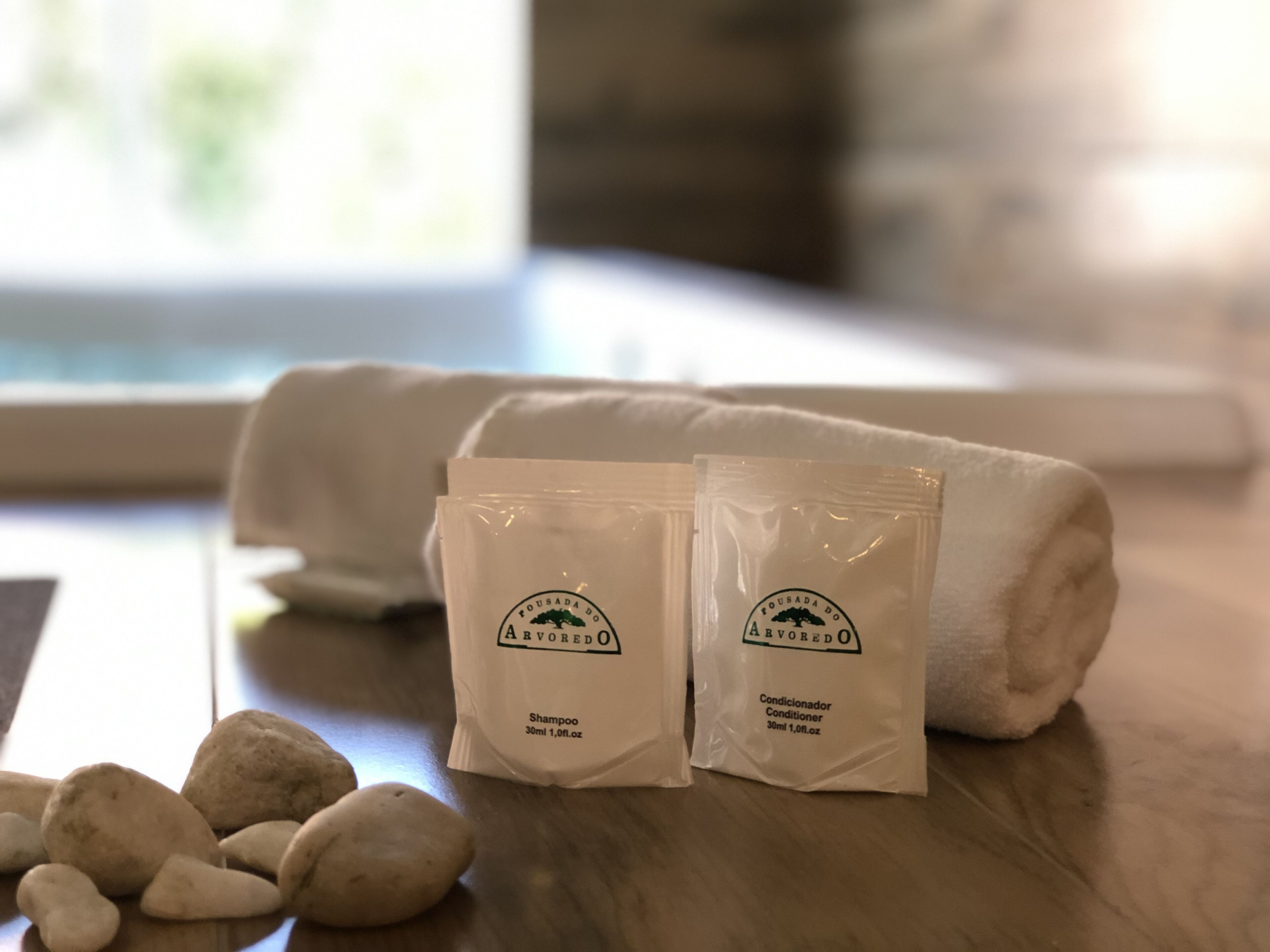 Dos pequeñas bolsas blancas, una con la etiqueta 'Fousada do Arvoredo Shampoo' y otra con 'Fousada do Arvoredo Conditioner', sobre una superficie de madera junto a una toalla blanca enrollada y algunas piedras blancas.