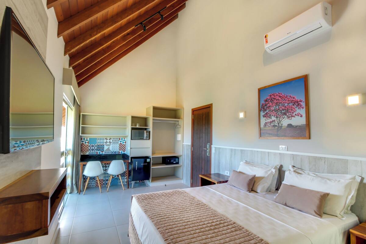 A bedroom with a bed, bedside tables, a desk with chairs, a wardrobe, and an air conditioning unit.