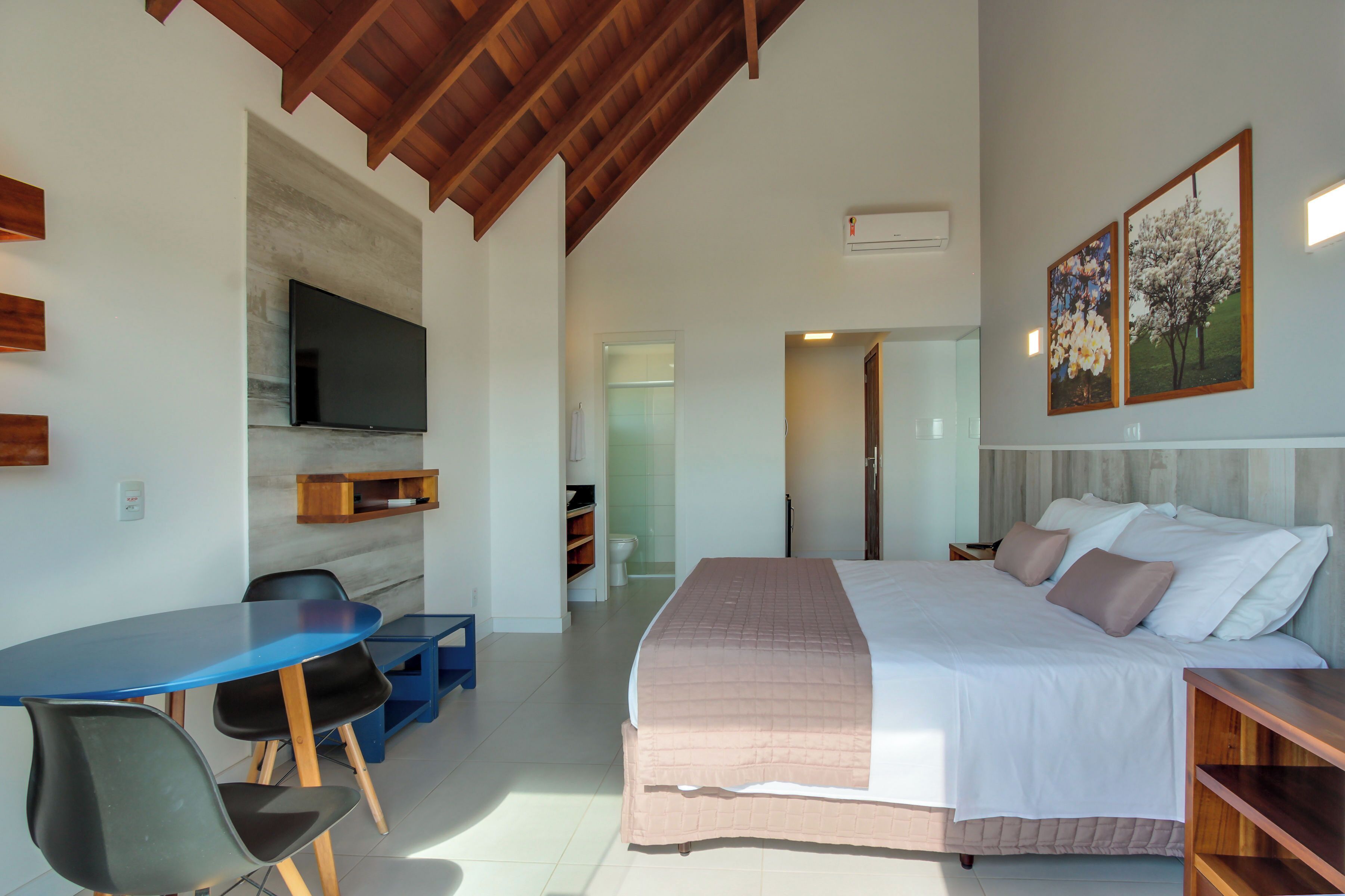 Een moderne hotelkamer met een groot bed, een flatscreen televisie aan de muur, een zithoek met een blauwe tafel en stoelen, en een houten plafond.