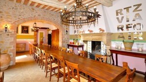 Una larga mesa de madera para comer con sillas, una araña, paredes de piedra y una chimenea.