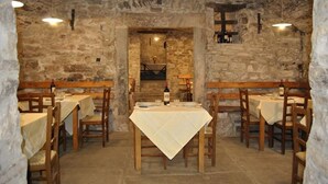 A stone-walled dining area with wooden tables and benches, a central table with a bottle of wine, and a small shelf with bottles.