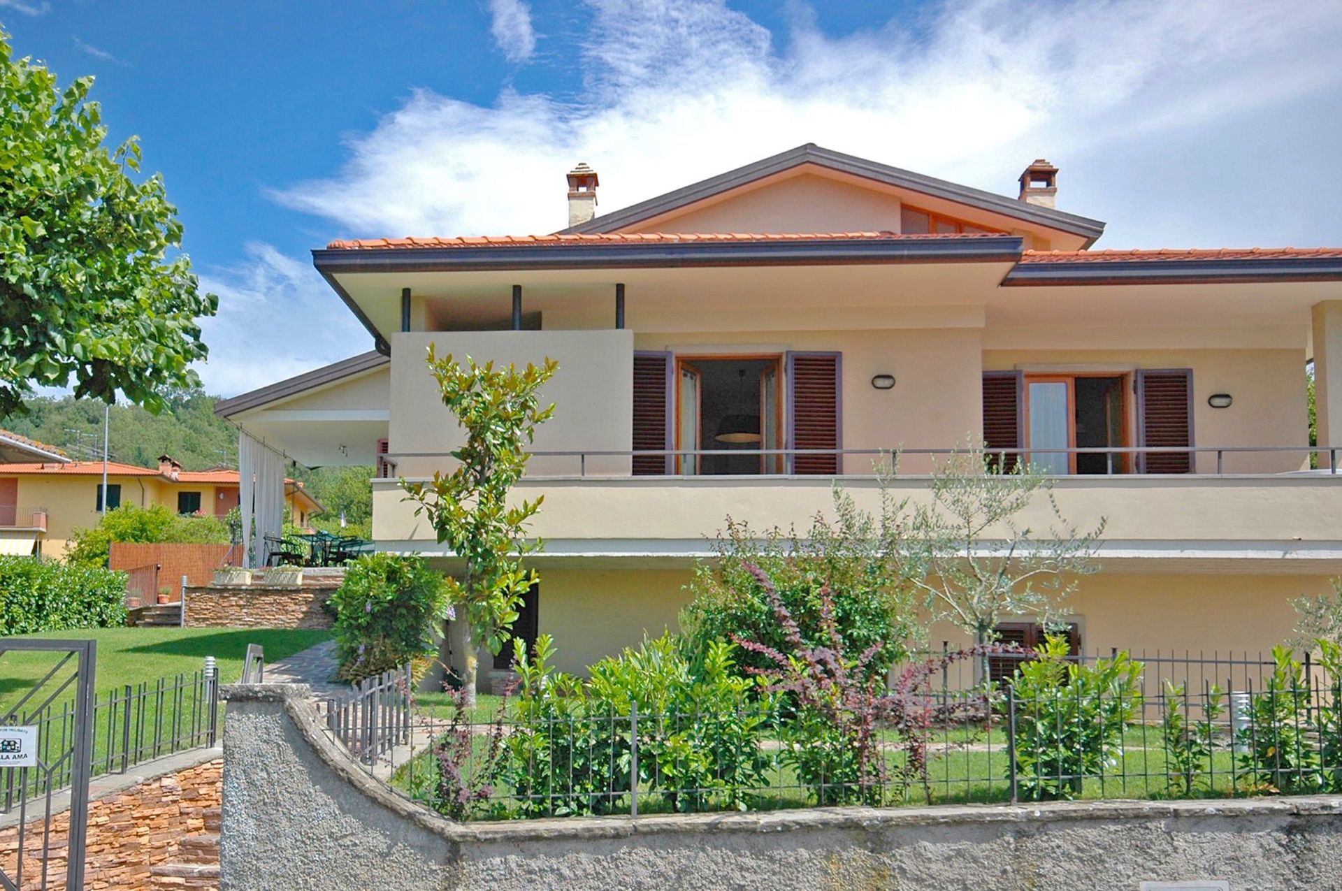 A two-story residential building with a balcony, a stone wall in front, and a clear sky.