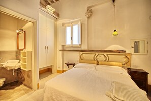 A bedroom with a large bed, a wooden headboard, a nightstand, a bedside table, a window, and a bathroom with a sink and mirror.