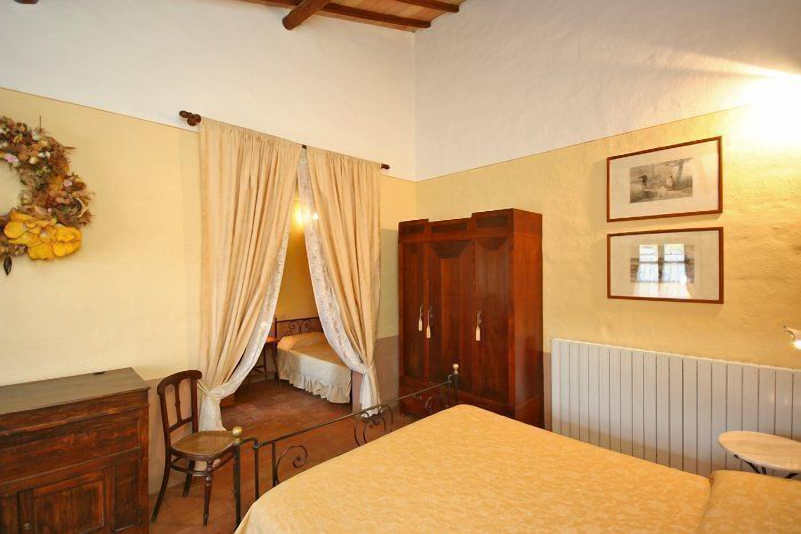 A bedroom with a bed, a wooden wardrobe, a chair, a small table, and a wall with two framed pictures.
