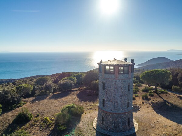 A stone tower with a circular base and a conical roof, surrounded by sparse vegetation and a clear view of the sea and distant hills.