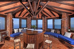 A circular wooden table surrounded by high-backed chairs, with a brick wall and large windows offering a view of the ocean. - Torre Rivolta (Orbetello)