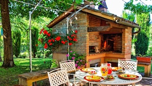 An outdoor dining area with a brick oven, white chairs, and a table set for a meal. - Villa San Savino (Monte San Savino)