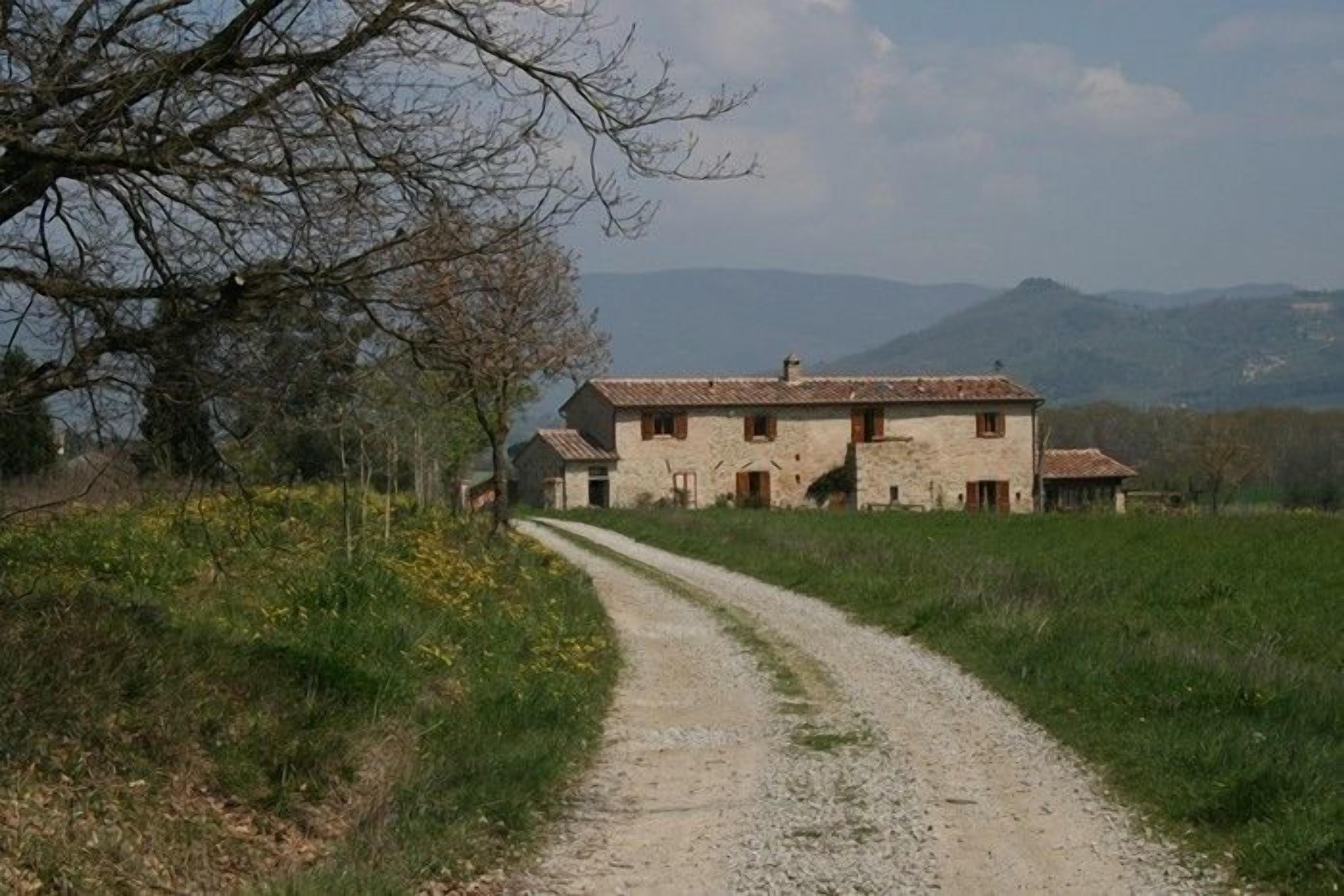 A rural road leading to a stone farmhouse with multiple buildings, surrounded by fields and mountains in the background.
