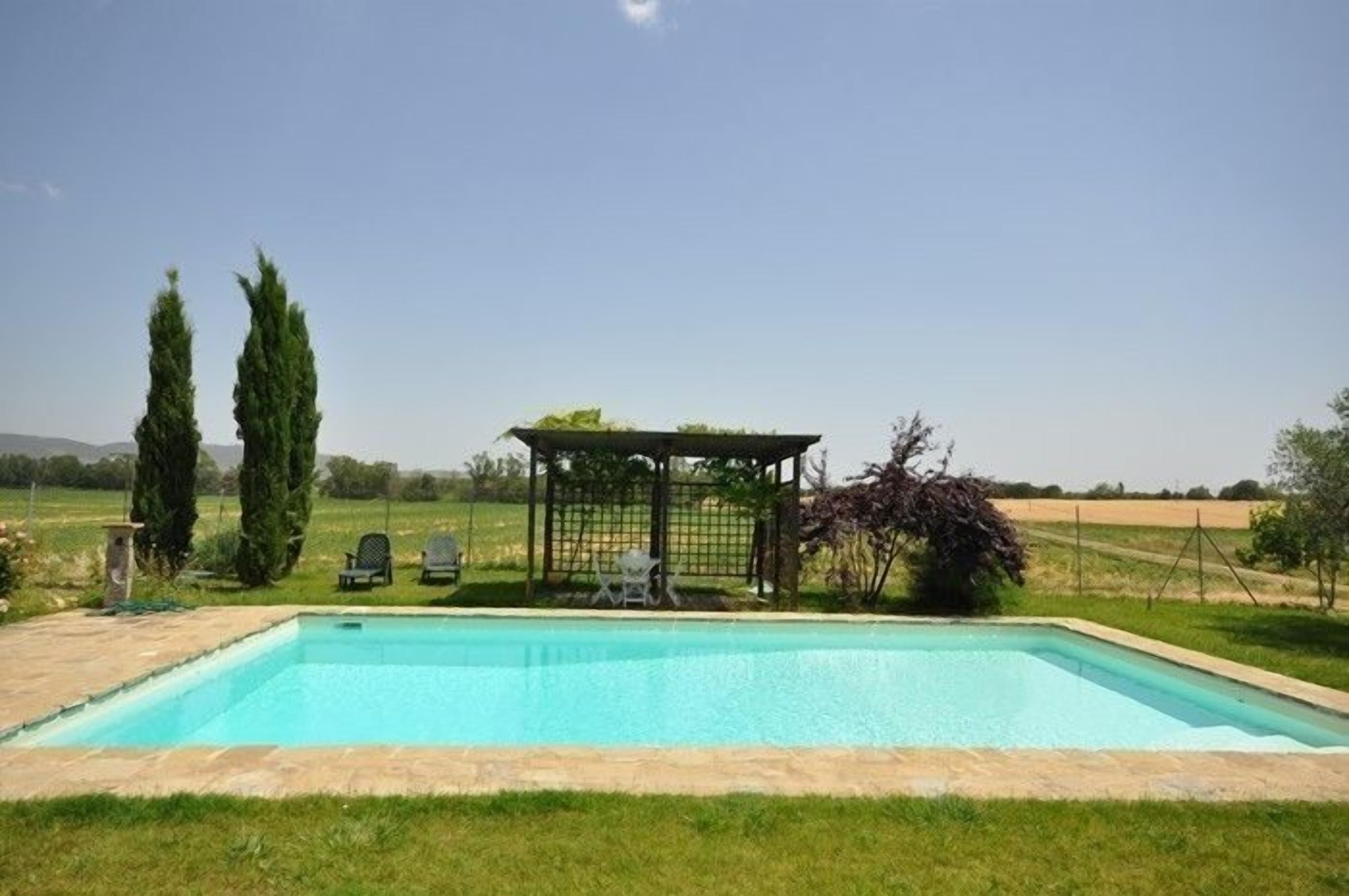 Una piscina rectangular con una pérgola y sillas al fondo.