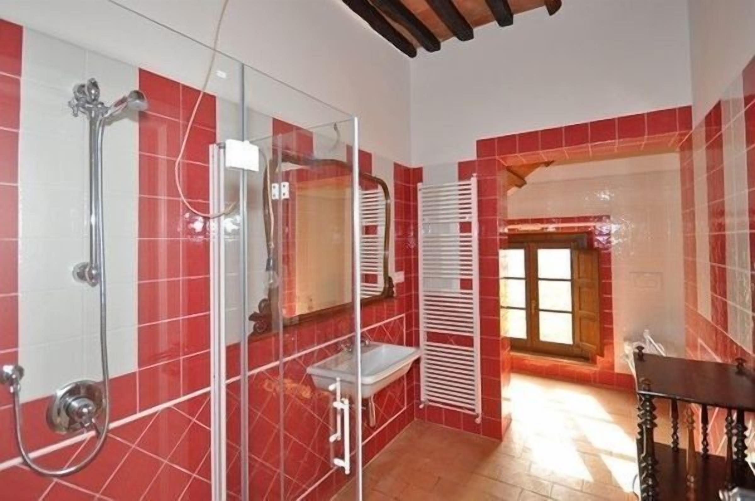 A bathroom with red tiled walls, a glass shower enclosure, a white sink, and a wooden bench.