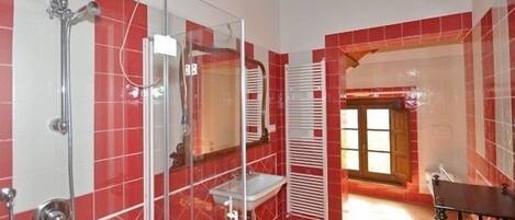 A bathroom with red tiled walls, a glass shower enclosure, a white sink, and a wooden bench.