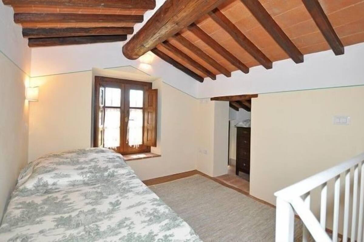 A bedroom with a bed, wooden ceiling beams, a window, and a door leading to another room.